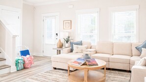 Una sala de estar con un sofá color beige, una mesa de centro con libros, una escalera y una puerta.