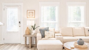 Una sala de estar con una puerta, una mesa con lámpara, un sofá y una ventana con persianas.