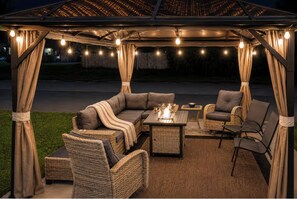 Un patio exterior con sofá, sillas y una mesa, todo bajo una zona techada con luces colgantes.