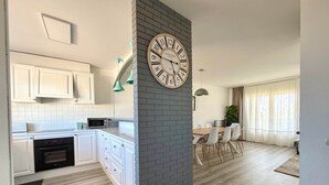 A modern kitchen with white cabinets, a microwave, and a built-in oven. There is a dining area with a table and chairs, and a clock on the wall. - V002 - Cargols de Mar 2 (Empuriabrava)