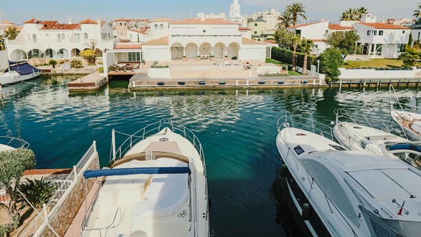 A marina with boats docked, a white building with arches, and a lighthouse in the background. - V002 - Cargols de Mar 2 (Empuriabrava)