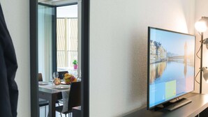 A modern hotel room with a flat-screen TV on a stand, a dining area with a table and chairs, and a view of a cityscape through the window. - Apartment  by Interhome (Lucerne)