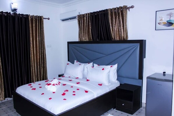 A neatly made bed with white linens and red rose petals, a dark headboard, and a small bedside table with a black drawer.