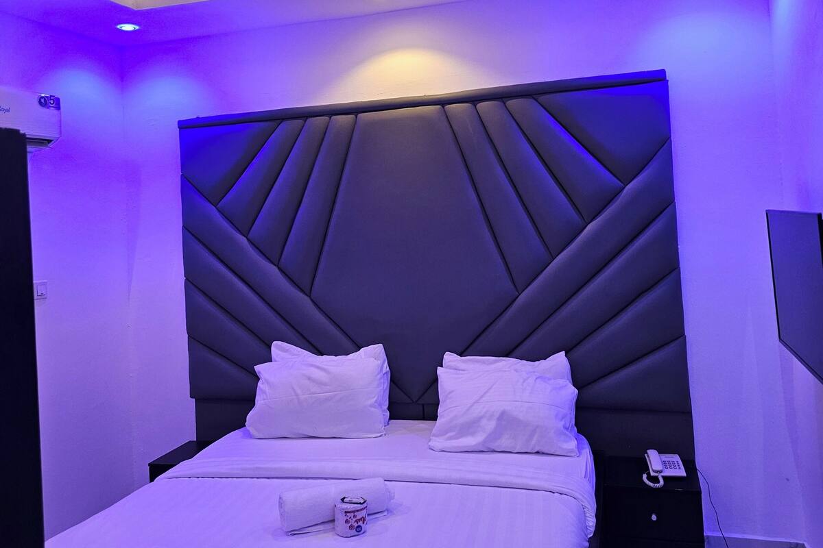 Une chambre moderne avec un plafond au design géométrique, un grand lit recouvert de linge de maison blanc et un téléphone intégré sur la table de chevet.