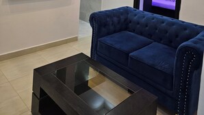 Ett modernt vardagsrum med en blå soffa, ett soffbord i glas, en kristallkrona och en väggmonterad tv som visar ett sovrum.