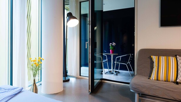 A modern hotel room with a bed, a sofa, a glass door leading to a balcony, and a vase with flowers. - LU Gletschergarten II-Allmend HITrental Apartment by Interhome (Lucerne)