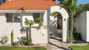 Une maison moderne avec un toit recouvert de tuiles rouges, un panneau à l’effigie du groupe U2 et un jardin bien entretenu où l’on peut admirer différentes espèces de cactus et de palmiers.