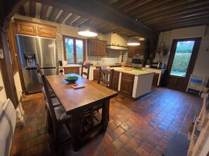 A kitchen with wooden cabinets, a stainless steel refrigerator, a dining table with chairs, and a large window. - Large Norman family home 5km from Deauville (Touques)