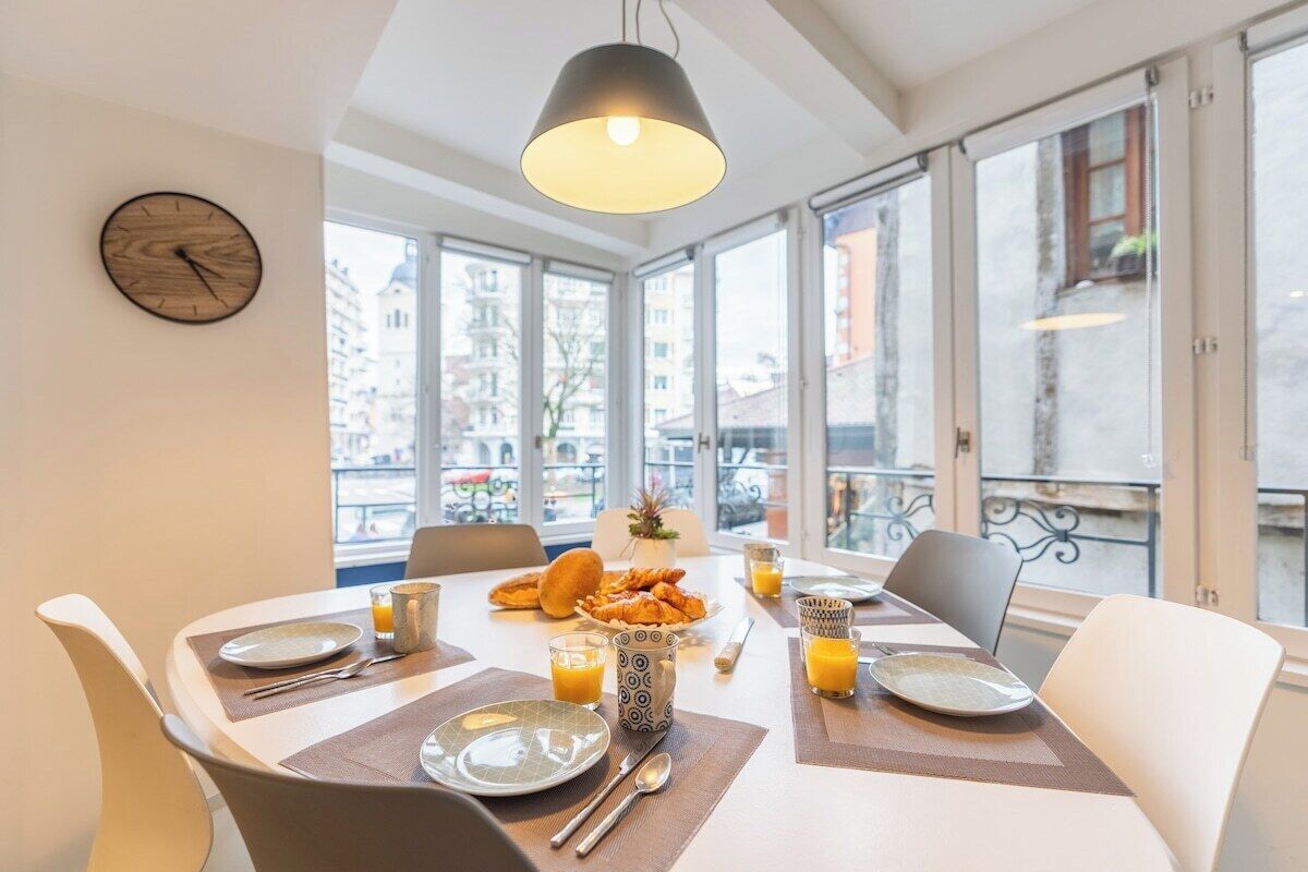 Une table de petit-déjeuner, avec des chaises, une horloge et une vue sur les bâtiments à travers de grandes fenêtres.