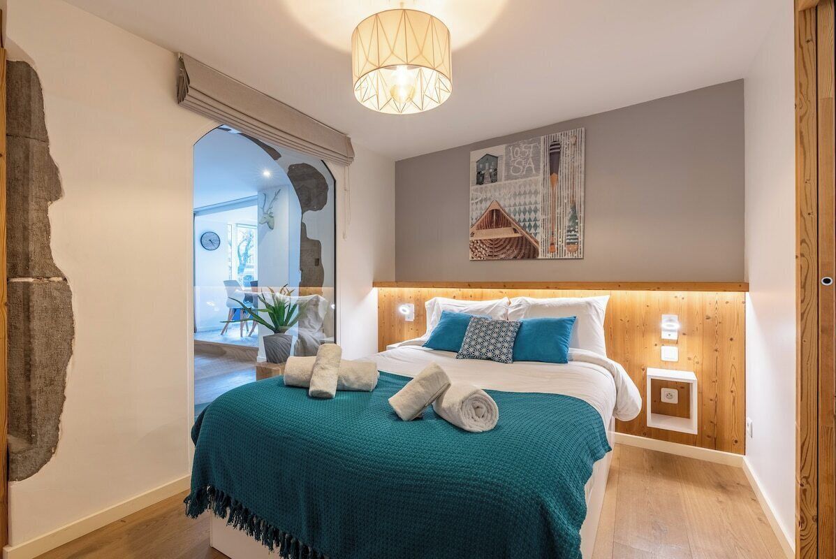 Une chambre à coucher avec un lit, une couverture turquoise, une tête de lit en bois, une lampe fixée au mur et un tableau.
