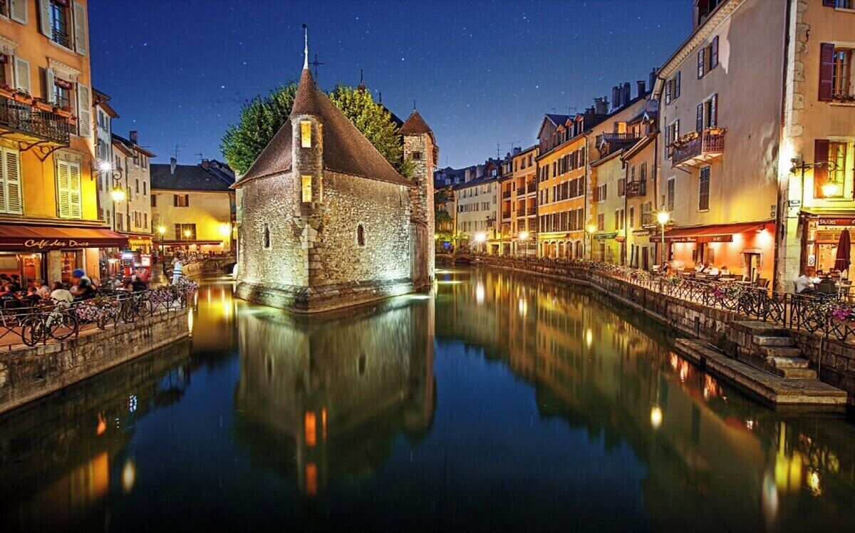 A historic stone building with a tower, reflecting in a calm water canal, surrounded by traditional European architecture.
