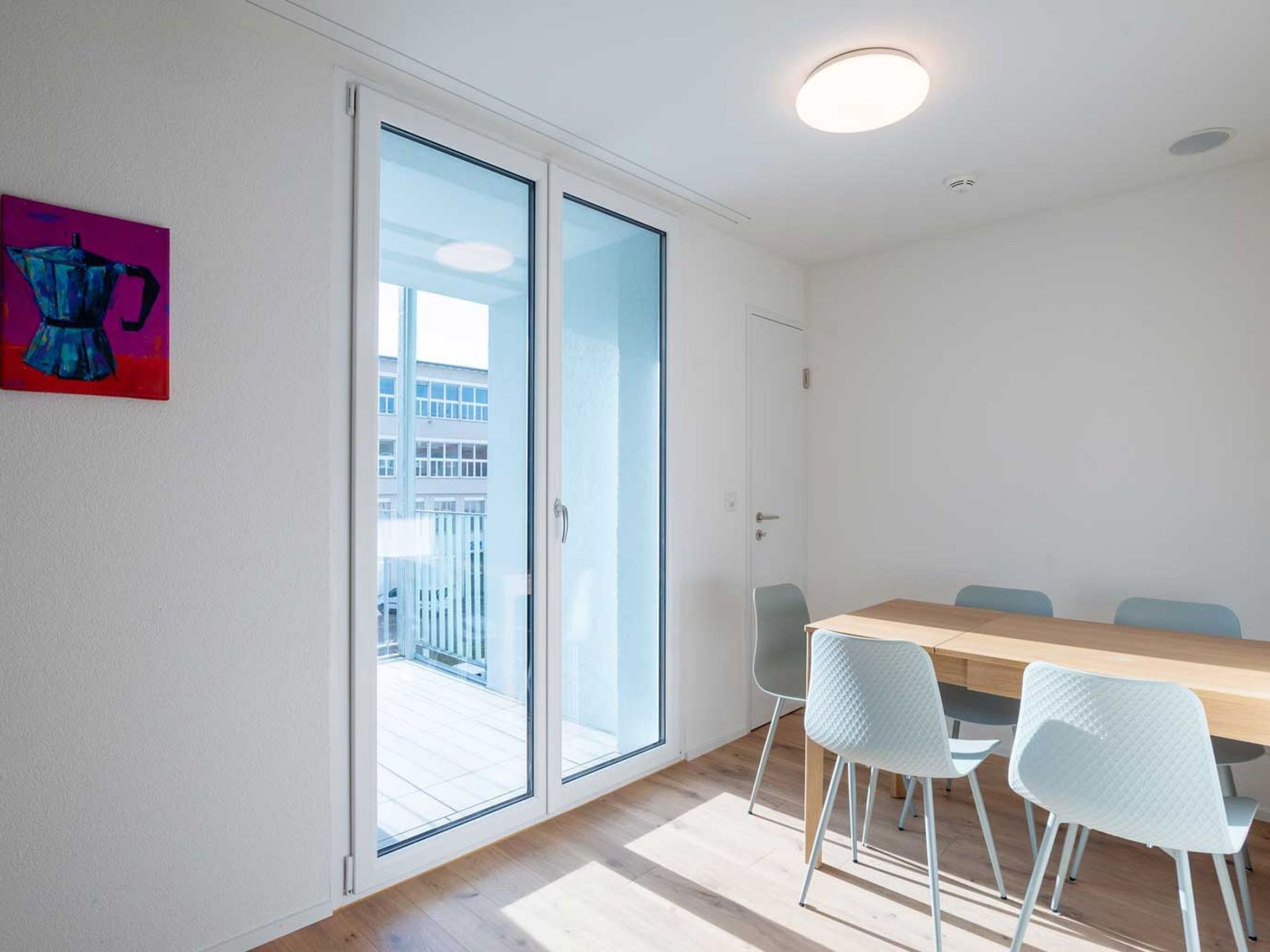 Una habitación con una mesa de madera y cuatro sillas, una puerta corrediza de vidrio que da a un balcón exterior y un cuadro pequeño en la pared.