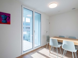 Una habitación con una mesa de madera y cuatro sillas, una puerta corrediza de vidrio que da a un balcón exterior y un cuadro pequeño en la pared.