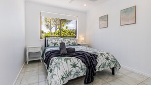 A bedroom with a bed, a nightstand, a window with a view of greenery, and two framed pictures on the wall. - Anchor and Palms boat parking, pet friendly air con free wifi - walk to beach & cafes (Agnes Water)