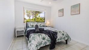 A bedroom with a bed, a nightstand, a window with a view of greenery, and two framed pictures on the wall. - Anchor and Palms boat parking, pet friendly air con free wifi - walk to beach & cafes (Agnes Water)