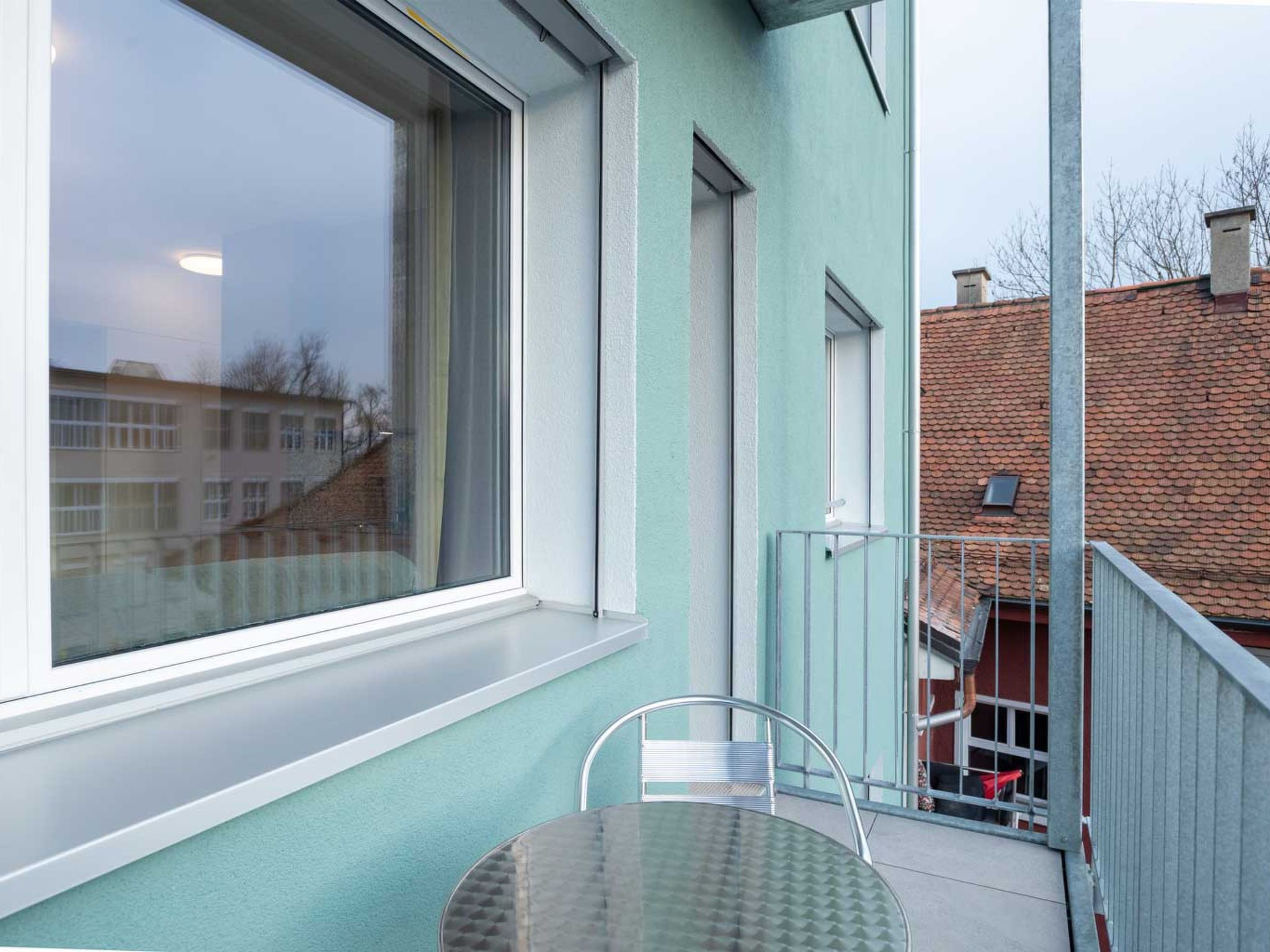 A balcony with a glass table and chairs, a large window, and a view of a building with a red roof.