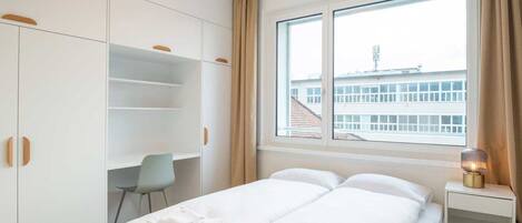 A bedroom with a bed, a desk, and a window with a view of buildings.