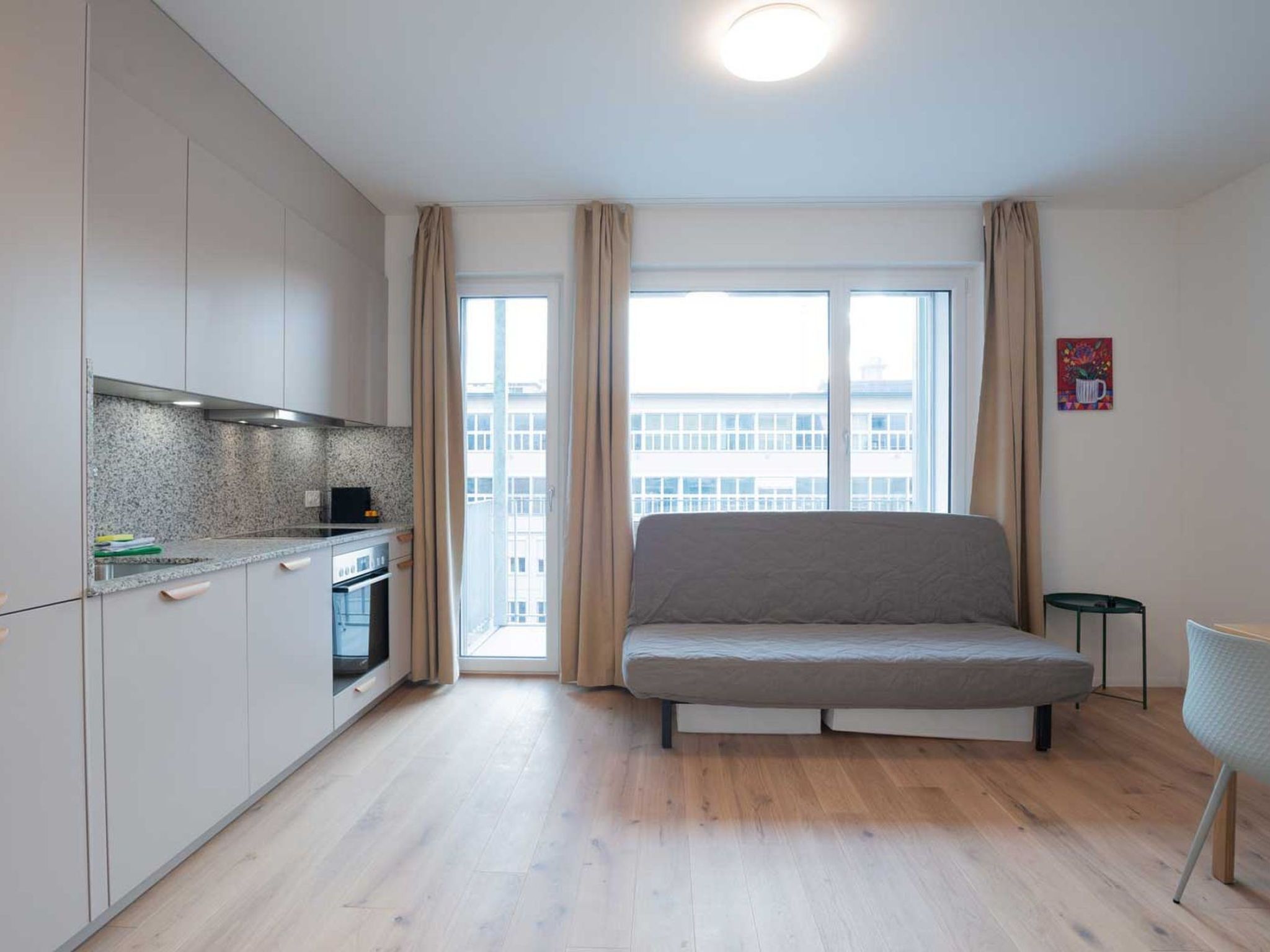 A modern apartment with a kitchenette, a sofa, and a view of a building through large windows.