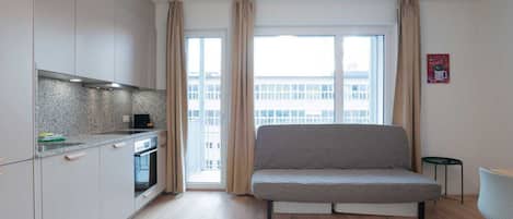 A modern apartment with a kitchenette, a sofa, and a view of a building through large windows.