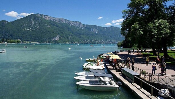 A lakeside view with boats docked, a promenade, and mountains in the background. - LakeView 1 - Premium Annecy - Veyrier du lac (Veyrier-du-Lac)