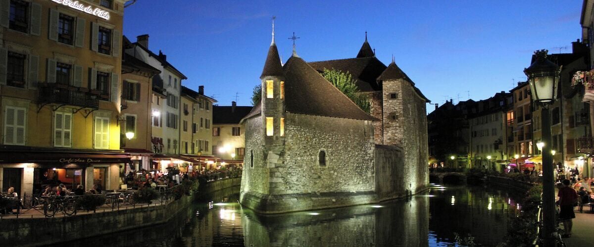 Ein historisches Steinhaus mit einem Turm, gelegen an einem Kanal mit einer Brücke im Hintergrund.