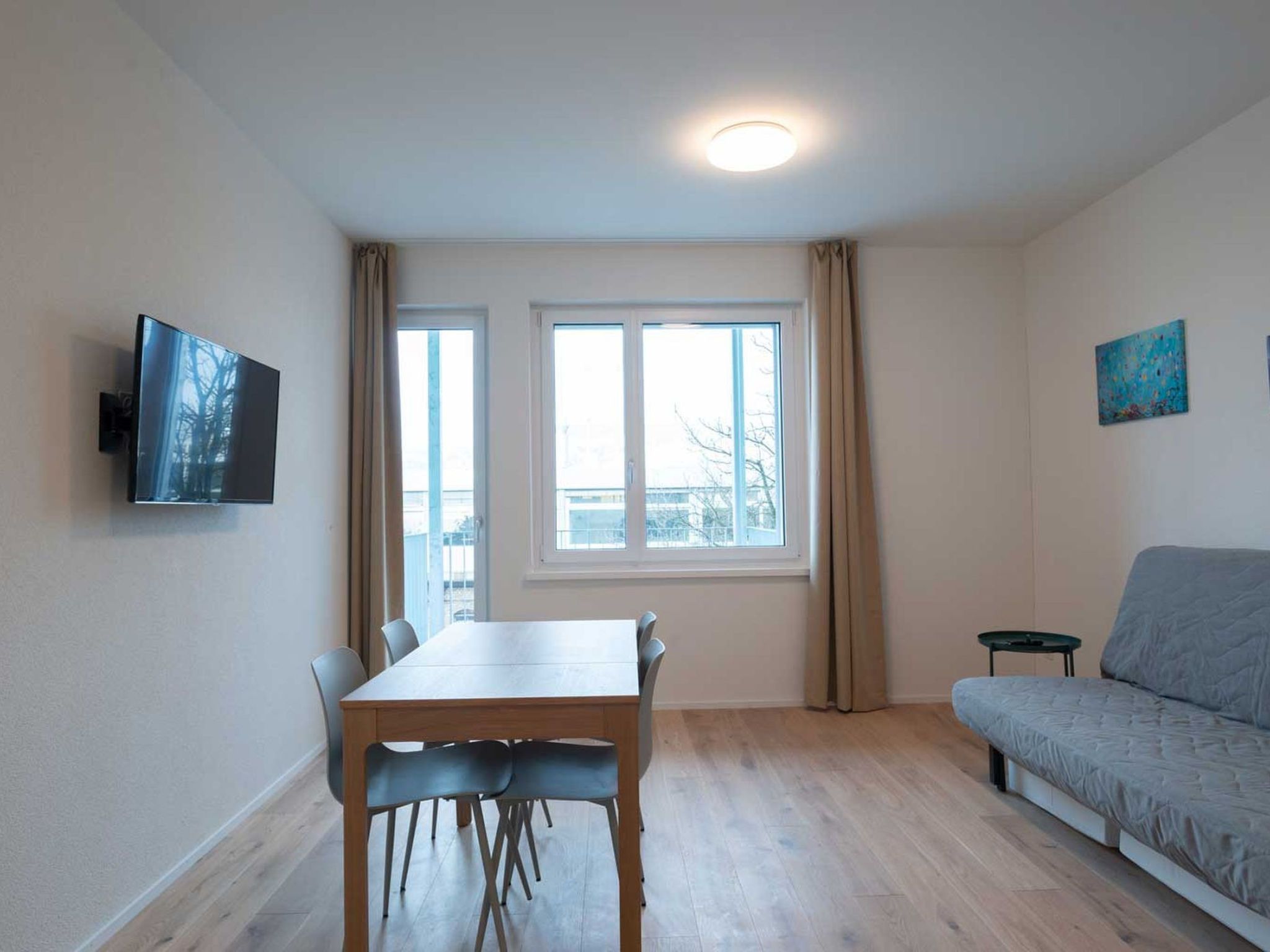 A room with a dining table, chairs, a sofa, a TV mounted on the wall, and two windows with curtains.