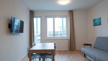 A room with a dining table, chairs, a sofa, a TV mounted on the wall, and two windows with curtains.