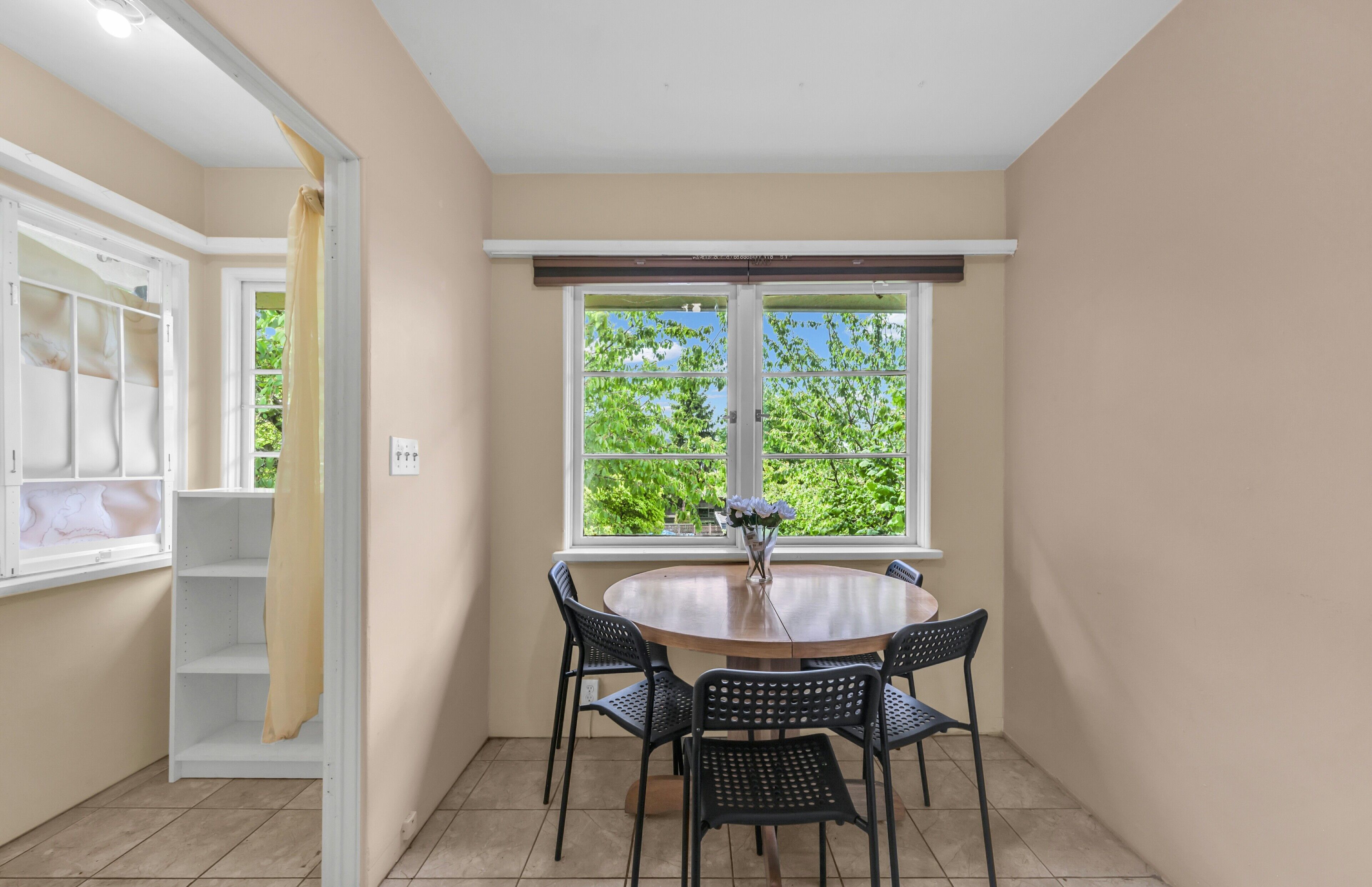 A room with a round table, four chairs, a window with a view of greenery, and a white door with a curtain.