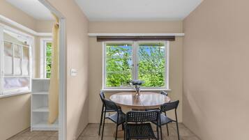 A room with a round table, four chairs, a window with a view of greenery, and a white door with a curtain.