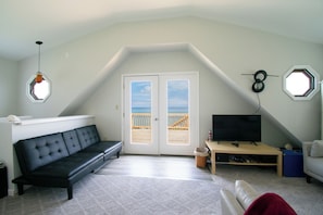Ein Zimmer mit einer schwarzen Couch, einem Fernseher auf einem Holzboden, einem kleinen MĂŒlleimer und Blick auf den Strand durch eine TĂŒr.