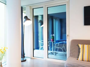 Ein modernes Wohnzimmer mit einer Couch und einem Tisch mit Stühlen, ein großes Fenster mit Vorhängen und eine Schiebetür zu einem Außenbereich mit Tisch und Stühlen.