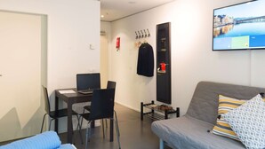 Ein modernes Wohnzimmer mit einer Couch, einem Schreibtisch mit Laptop, einem an der Wand befestigten Fernseher und einem Kleiderständer mit hängenden Gegenständen.