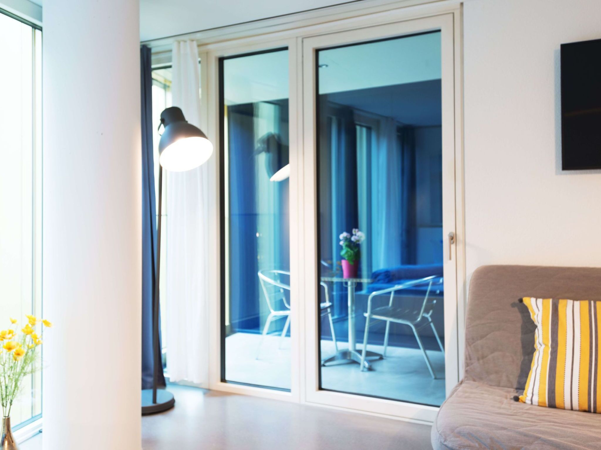 Ein modernes Wohnzimmer mit einer Couch und einem Tisch mit Stühlen, ein großes Fenster mit Vorhängen und eine Schiebetür zu einem Außenbereich mit Tisch und Stühlen.