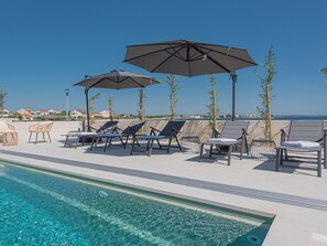 Een zwembadgedeelte met ligbedden en parasols, een helderblauwe lucht en uitzicht op de zee.