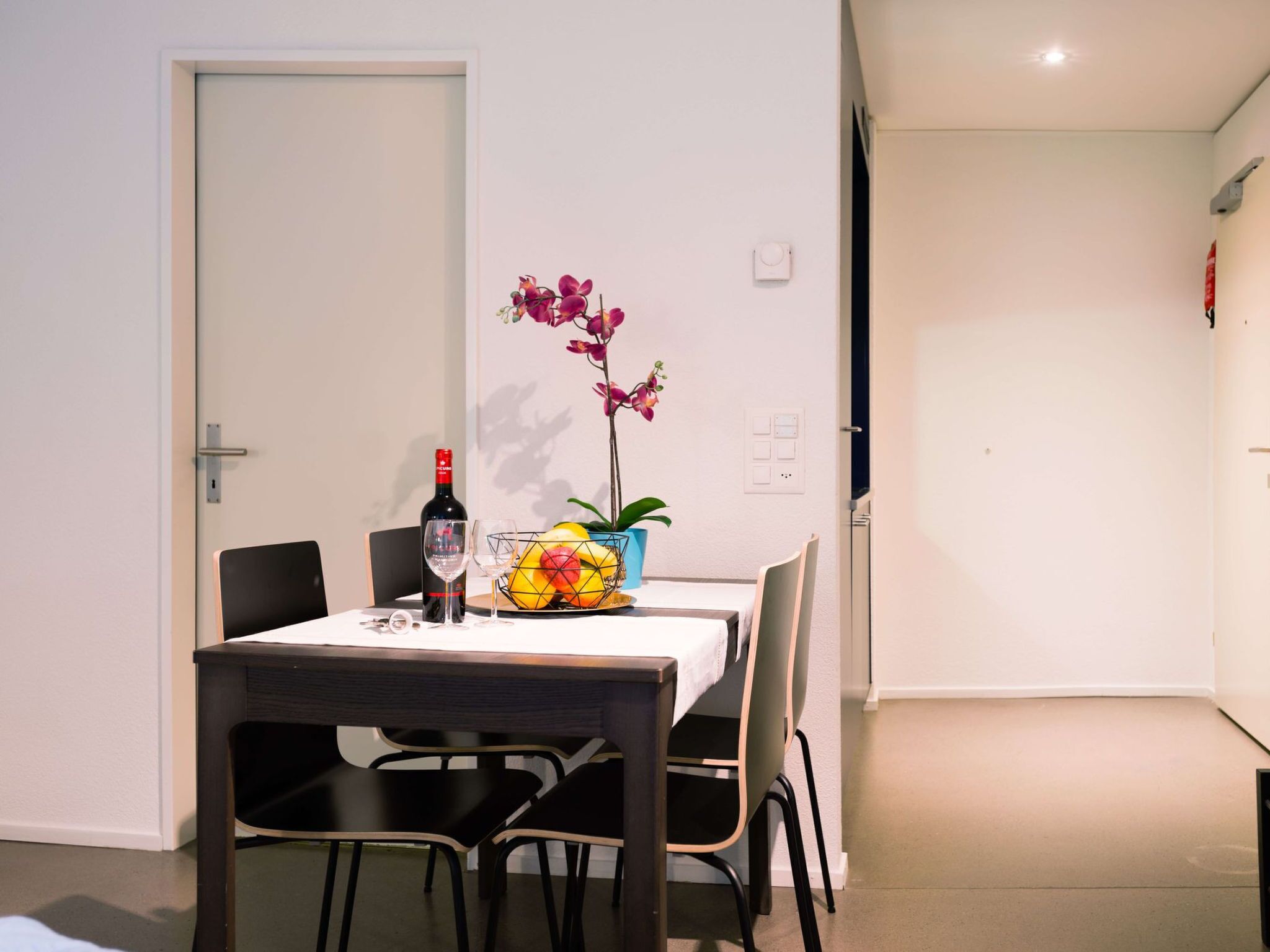 Un coin repas avec une table dressée pour deux personnes, une bouteille de vin et un vase rempli de fleurs.