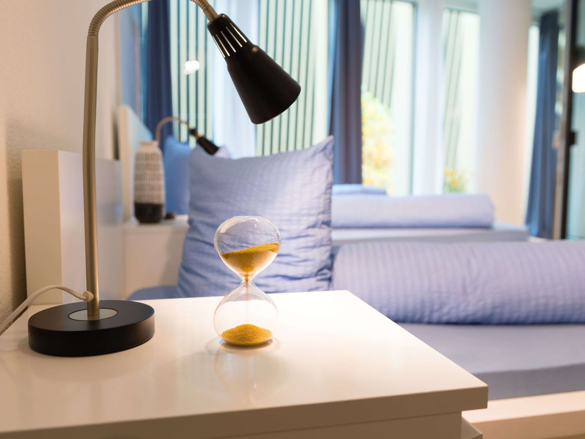 Ein Nachttisch mit Lampe, einer Sanduhr und ein Bett mit Kissen in einem Hotelzimmer.