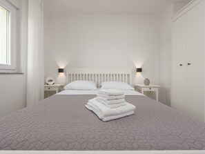 Un lit bien fait, recouvert d’une couette grise, avec des oreillers blancs et une pile de serviettes blanches. Il y a deux tables de chevet, chacune surmontée d’une lampe et d’un petit objet décoratif.