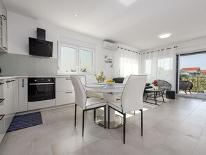 A modern kitchen with white cabinets, a built-in oven, and a dining area with a glass table and white chairs. - Apartment  by Interhome (Nin)