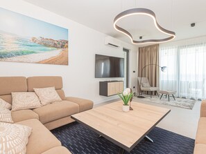 Un salon moderne comprenant un canapé, une table basse, un téléviseur à écran plat, un fauteuil et un tableau représentant un paysage côtier.