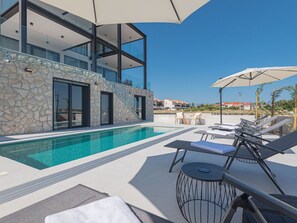 Un espace piscine moderne avec des chaises longues et des parasols, des murs en pierre et un ciel d’un bleu éclatant.