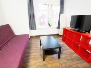 A room with a wooden floor, a red sofa, a black table, a red entertainment unit with a TV, and a window with curtains. - Apartment  by Interhome (Zurich)