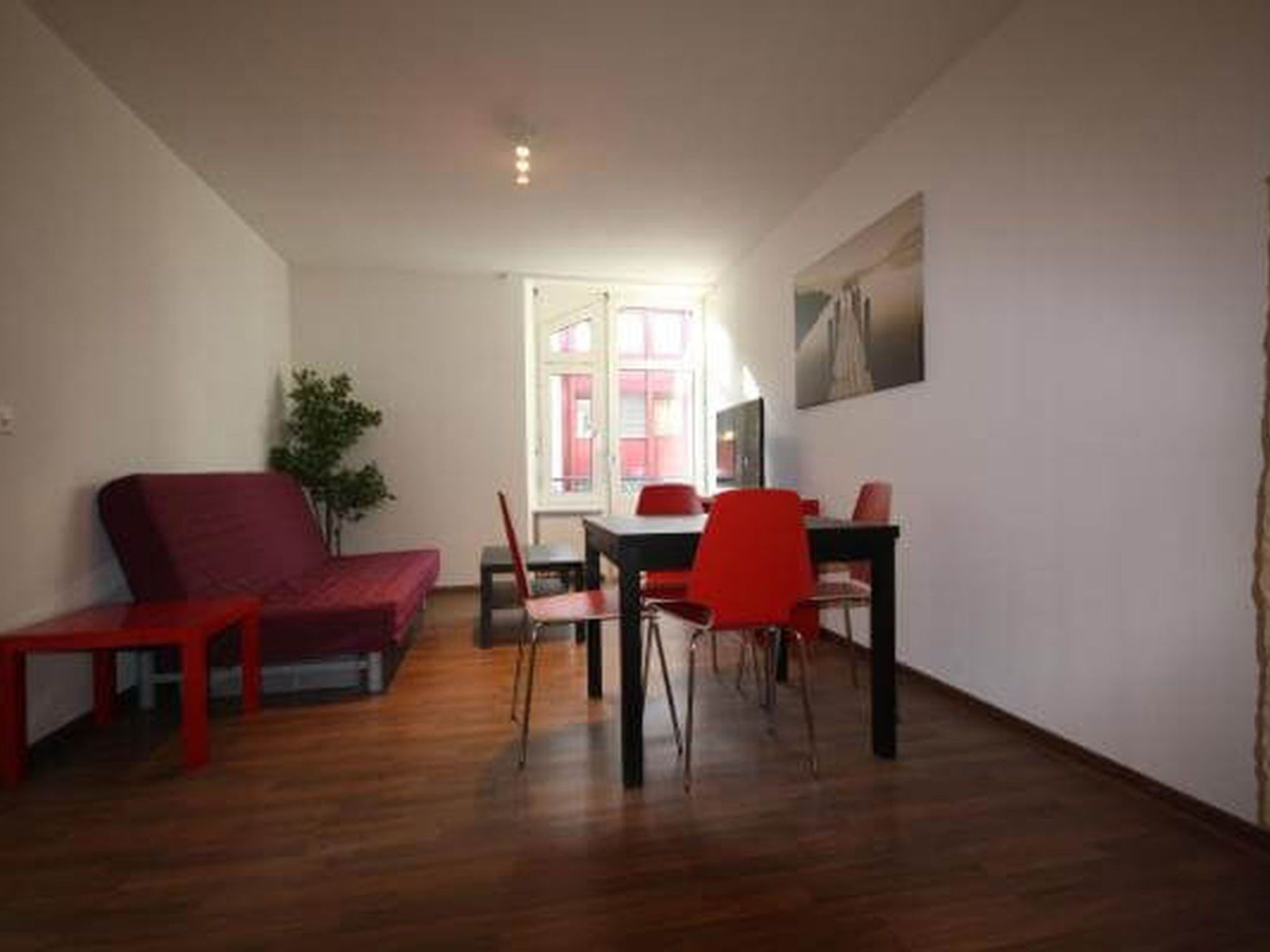A room with a red sofa, a dining table with chairs, a potted plant, and a window.
