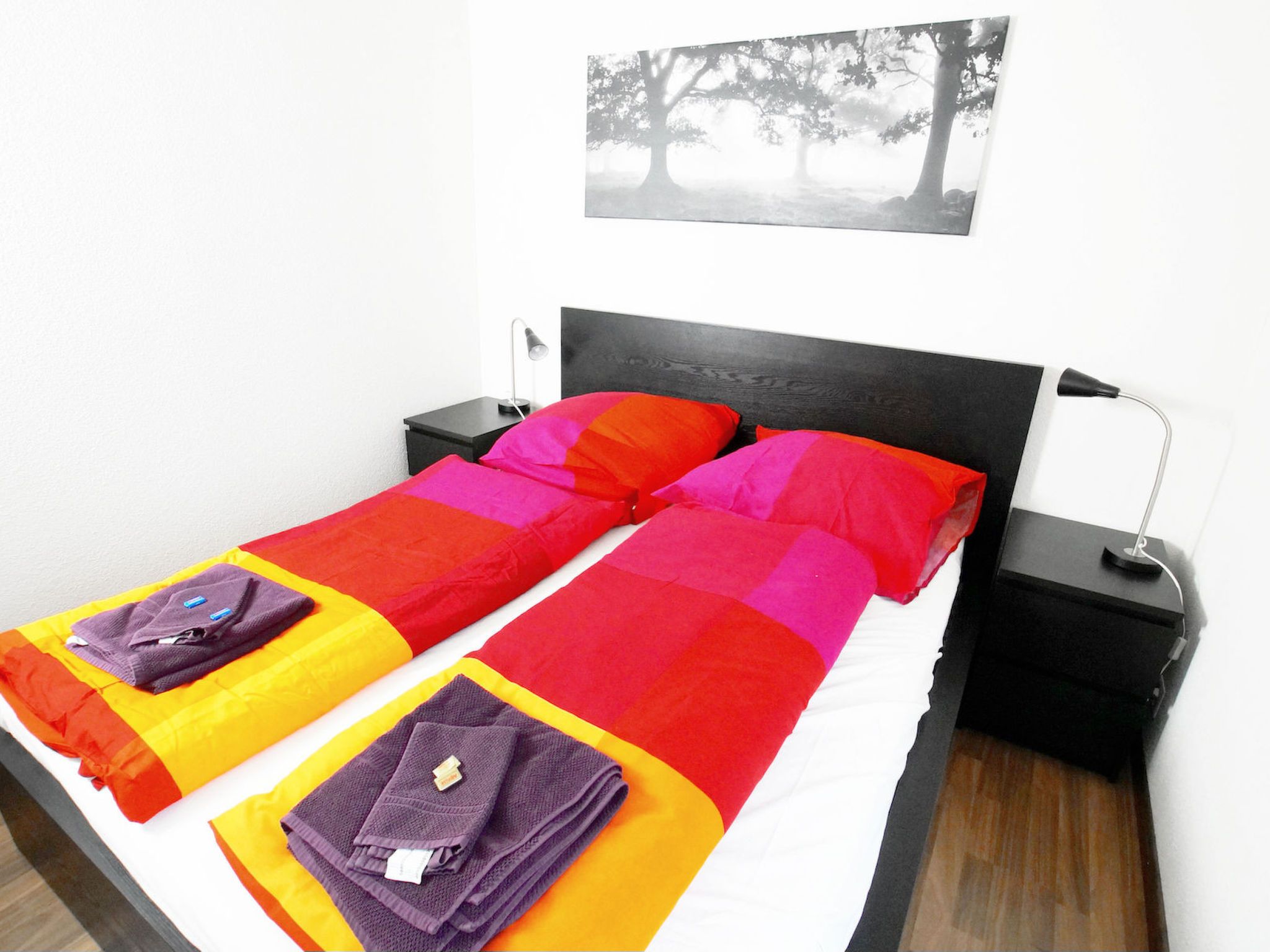 A double bed with a colorful bedspread and two pillows, a bedside table with a lamp, and a wall-mounted picture.