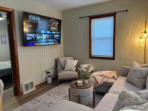 Una sala de estar con un televisor de pantalla plana montado en la pared, un sofá, una mesa de centro y una ventana con persianas.