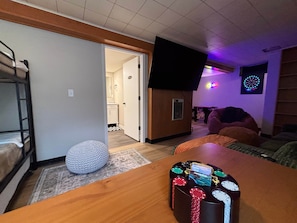 Una habitación con una cama litera, un pouf redondo, una mesa de madera con un portalápices para fichas de póker, un televisor montado en la pared y un dardo.