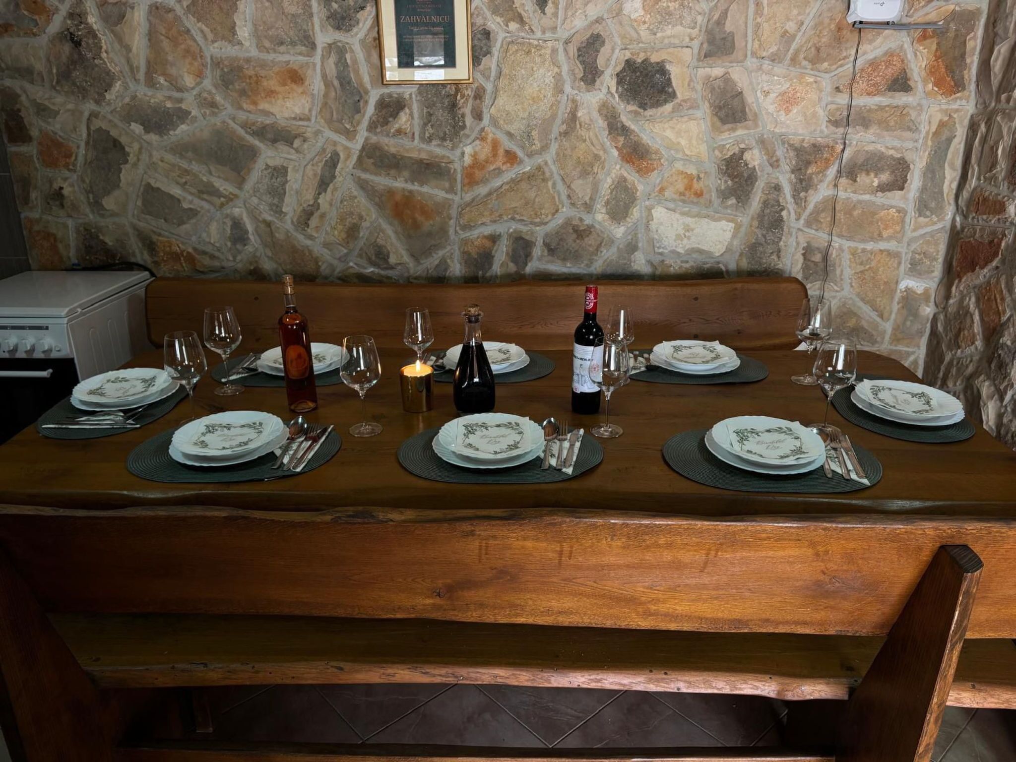 Una pared de piedra con un certificado enmarcado, una mesa de madera dispuesta para comer con platos, vasos y cubiertos, y un banco al fondo.
