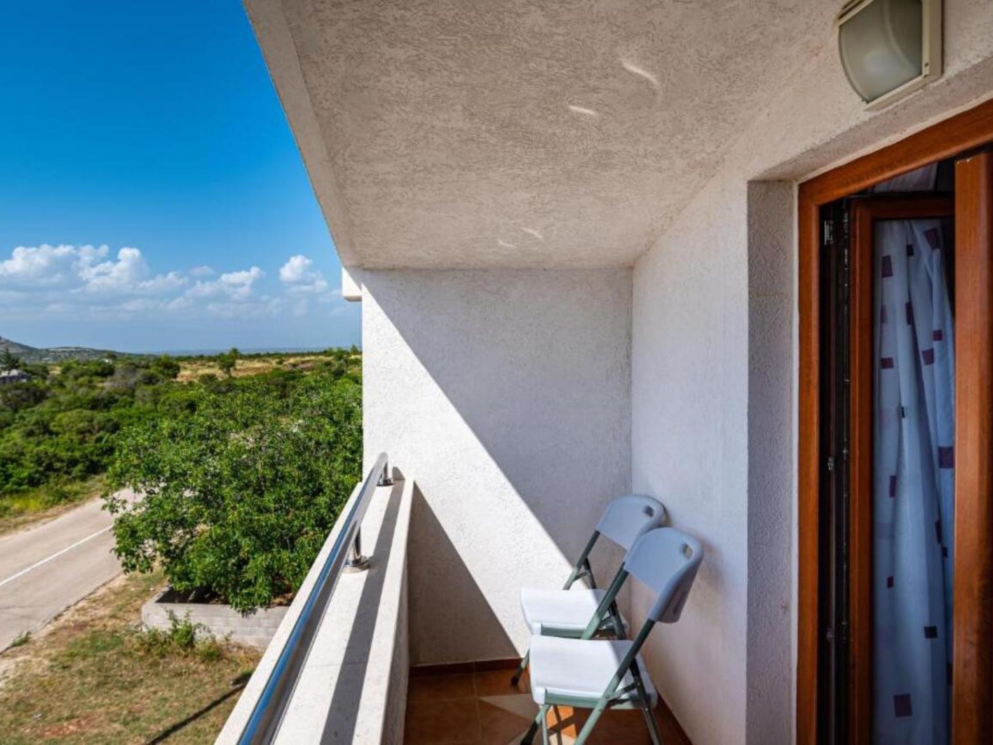 Un balcon donnant sur une route et sur de la verdure, une chaise blanche et une porte en bois avec un rideau.