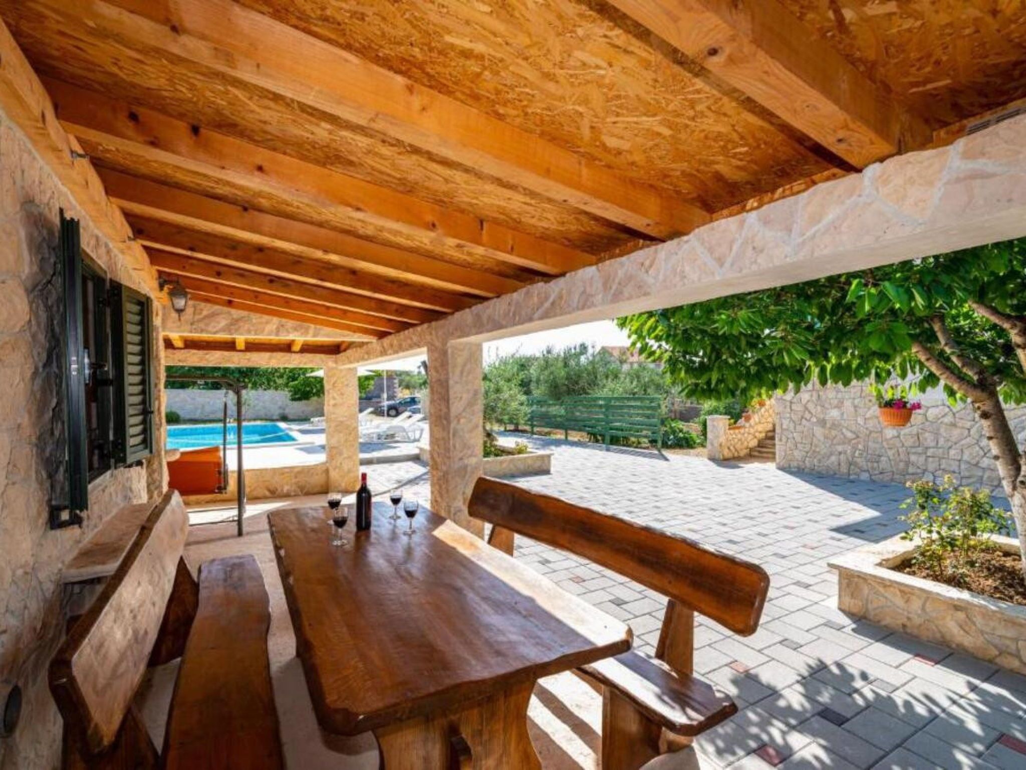 An outdoor patio with wooden benches and a table, stone walls, and a view of a swimming pool.