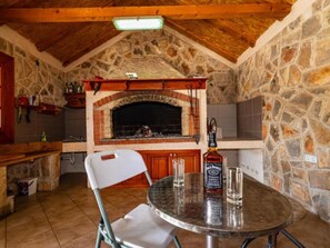 A stone fireplace with a brick arch, a metal table with glasses and a bottle, and a white folding chair in a room with wooden beams and stone walls. - Apartment Tonka 3+2 by Interhome (Donje Rastane)