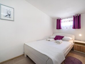 Ein ordentlich bezogenes Bett mit weißen Bettwäsche und einem violetten Kopfkissen, ein Nachttisch mit einer Lampe und ein Fenster mit violetten Vorhängen.